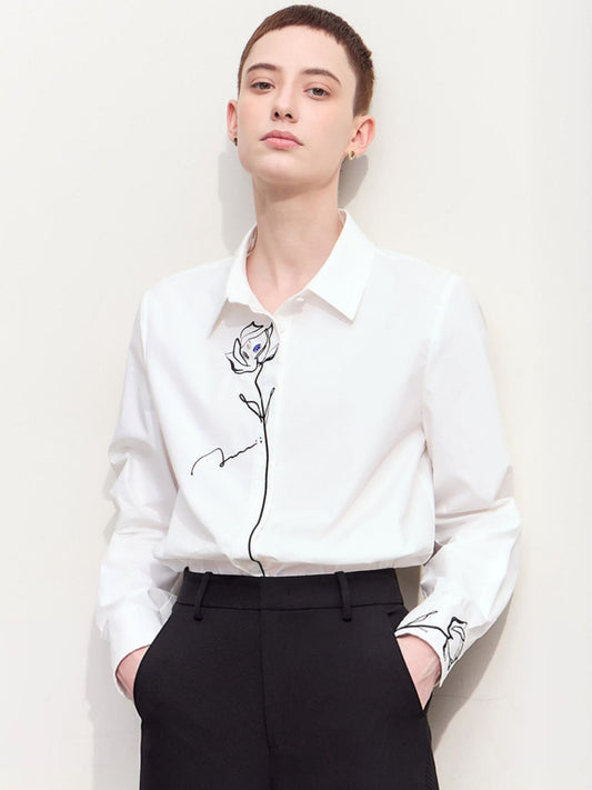 Hvid skjorte med sort blomsterbroderi, lange ærmer og klassisk krave, bæres med sorte bukser.