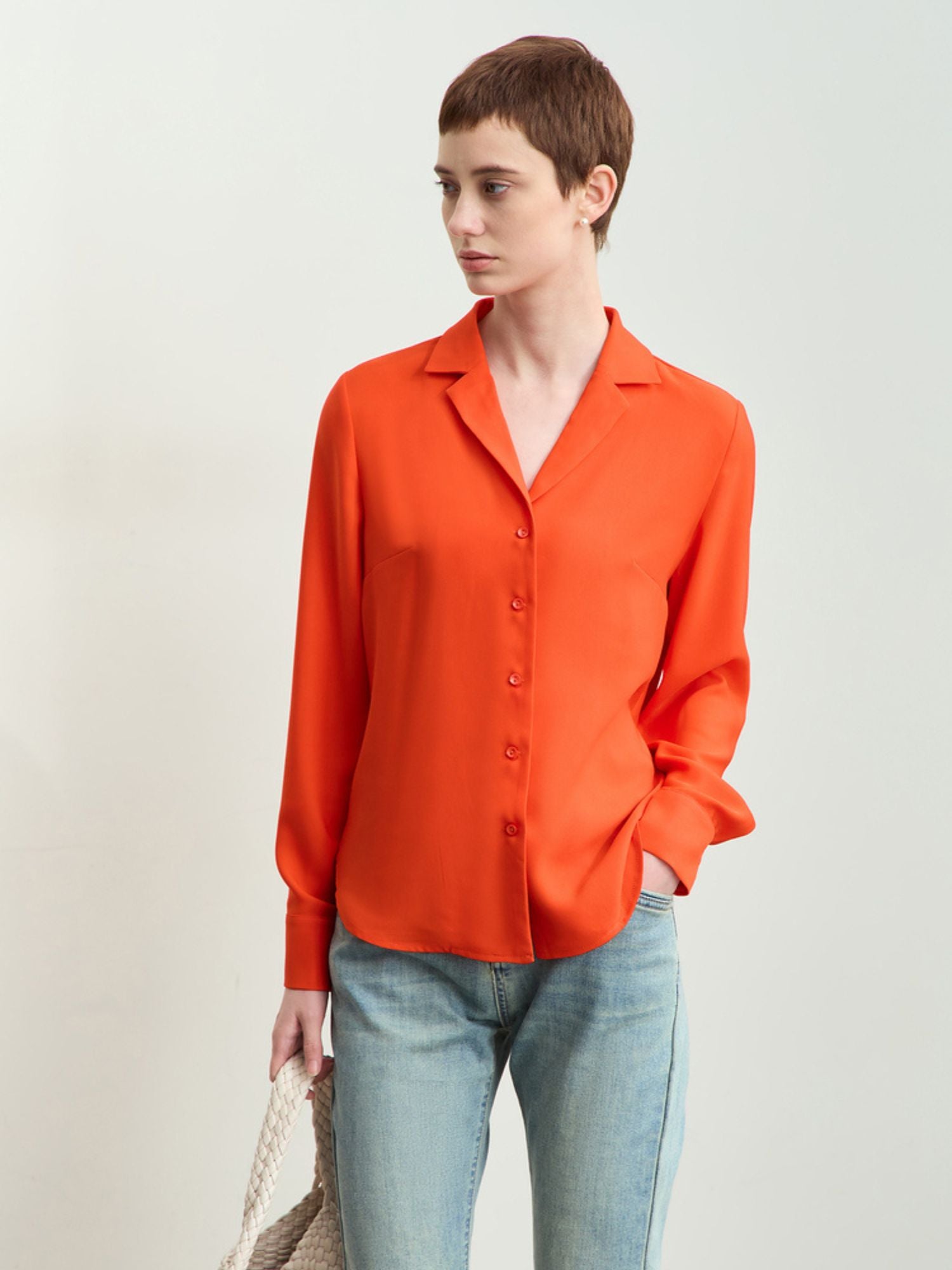 Kvinde iført rød-orange skjorte med krave og blå jeans, står med taske i hånden.