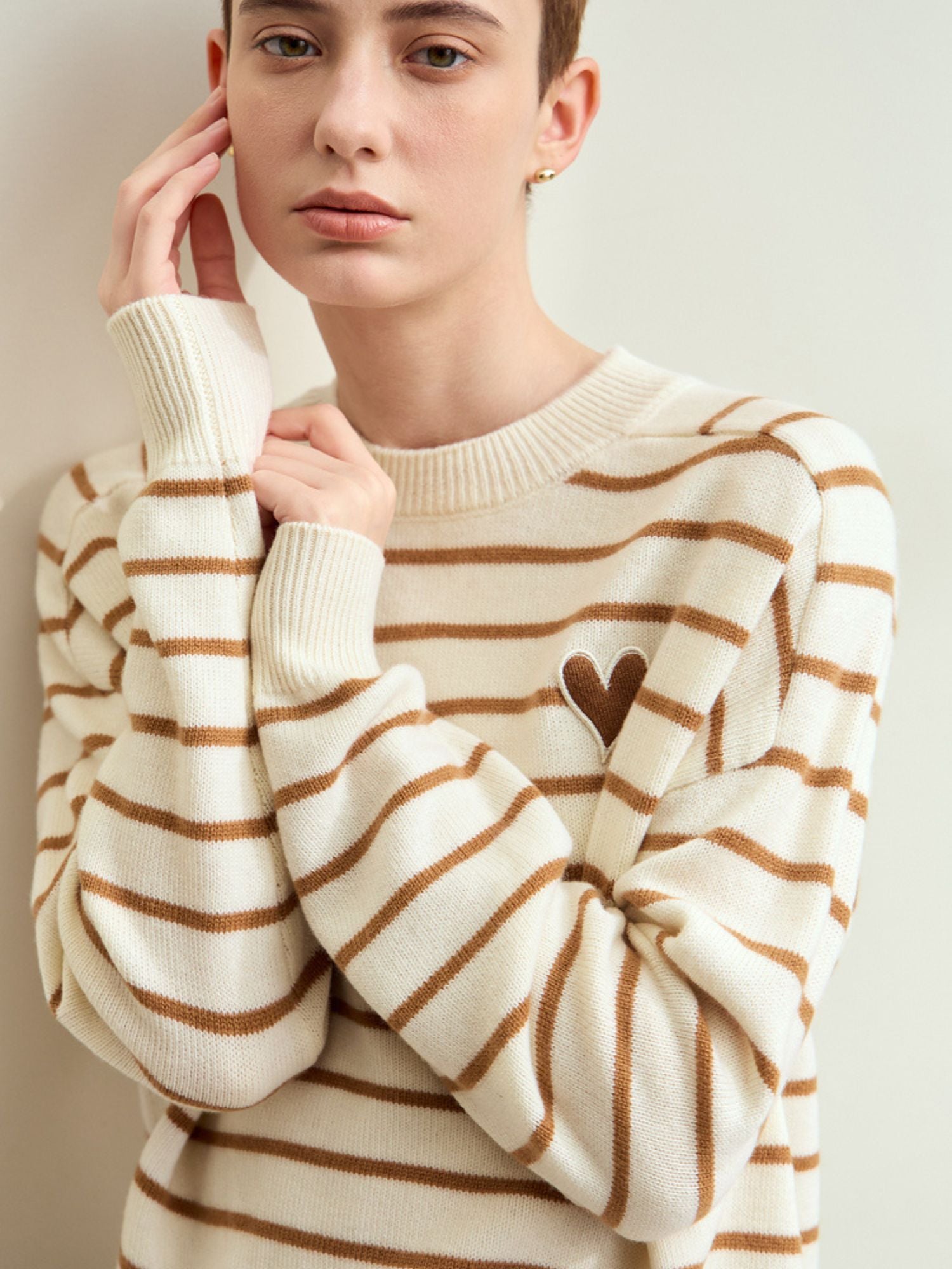 Model iført beige stribet sweater med hænderne mod ansigtet.