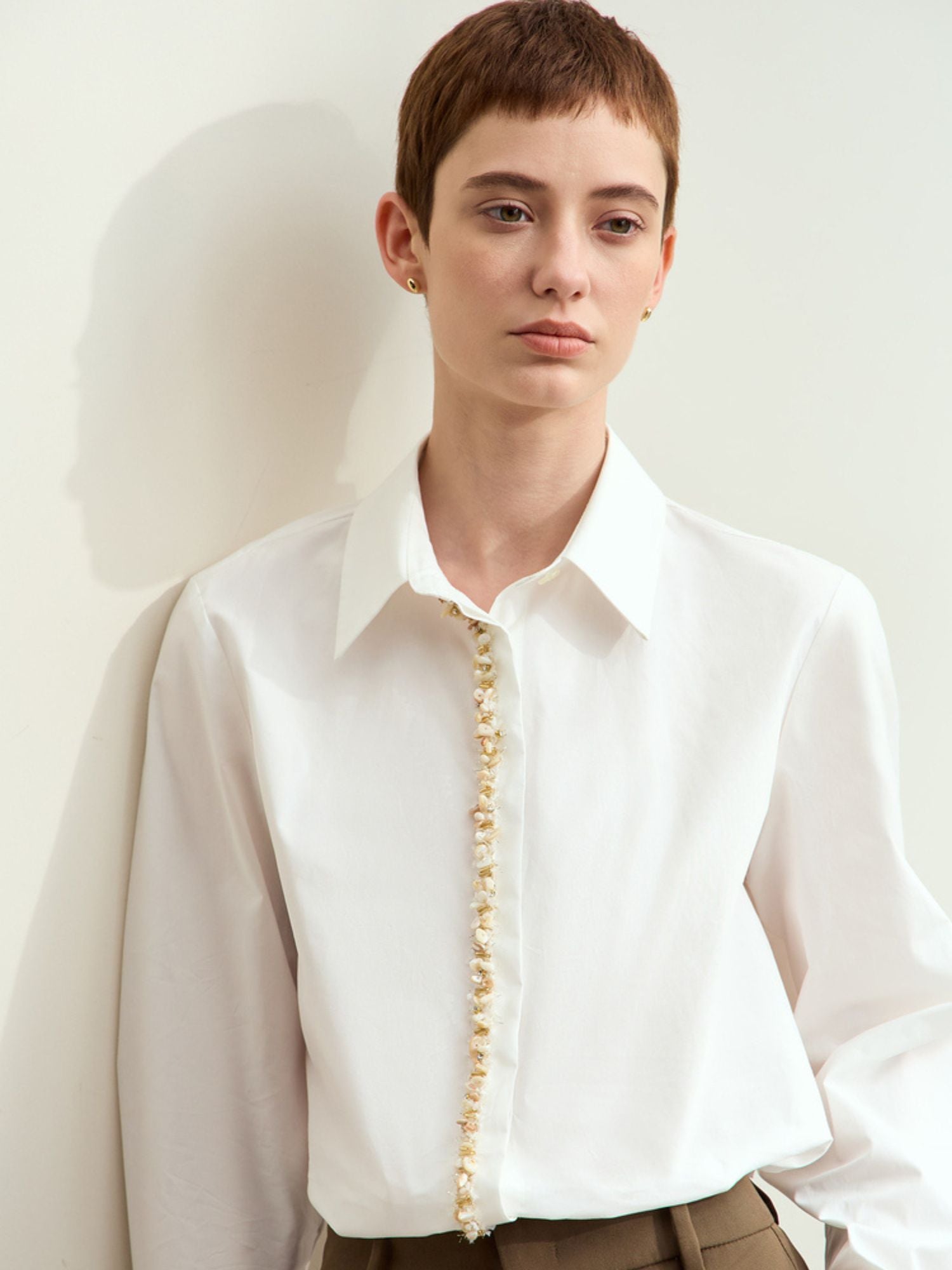 Skrå vinkel af model i hvid skjorte og brun nederdel, står op ad væg.